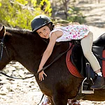 sweet beautiful young girl 7 or 8 years old riding pony horse hugging and smiling happy wearing safety jockey helmet posing outdoors on countryside in summer holiday