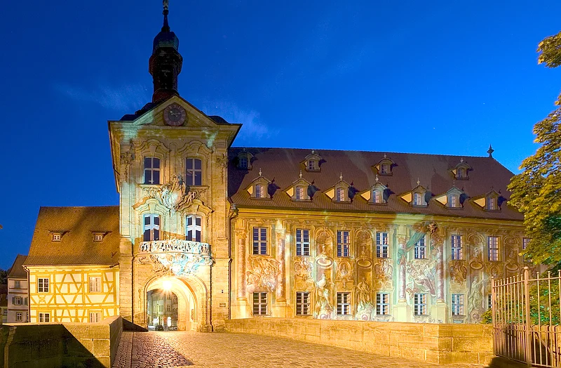 Bereits im frühen 11. Jahrhundert führte hier eine Brücke über den Fluss. Das in der Mitte des 14. Jahrhunderts gebaute Rathaus wurde nach einer Brandkatastrophe 1440 von den Bamberger Bürgern neu errichtet. Michael Küchel gestaltete es 1744-1756 im Stil des Barock und Rokoko um. Neben der wundervollen Architektur bestechen besonders die Fresken durch ihre Plastizität.